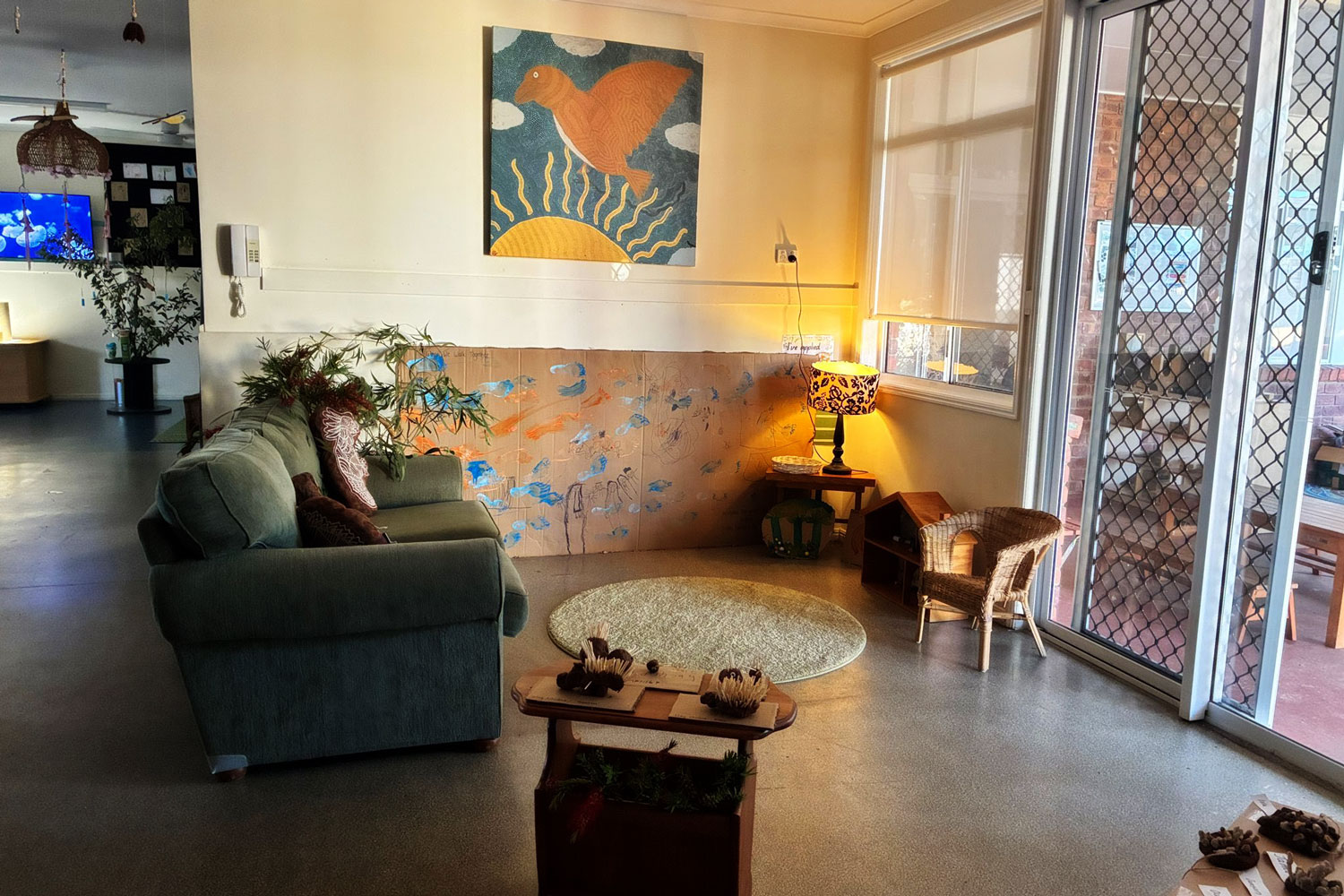 A photograph of an indoor kindergarten space. There is a green couch, a small wicker chair, a rug and a lamp on. The walls are adorned with children’s artwork