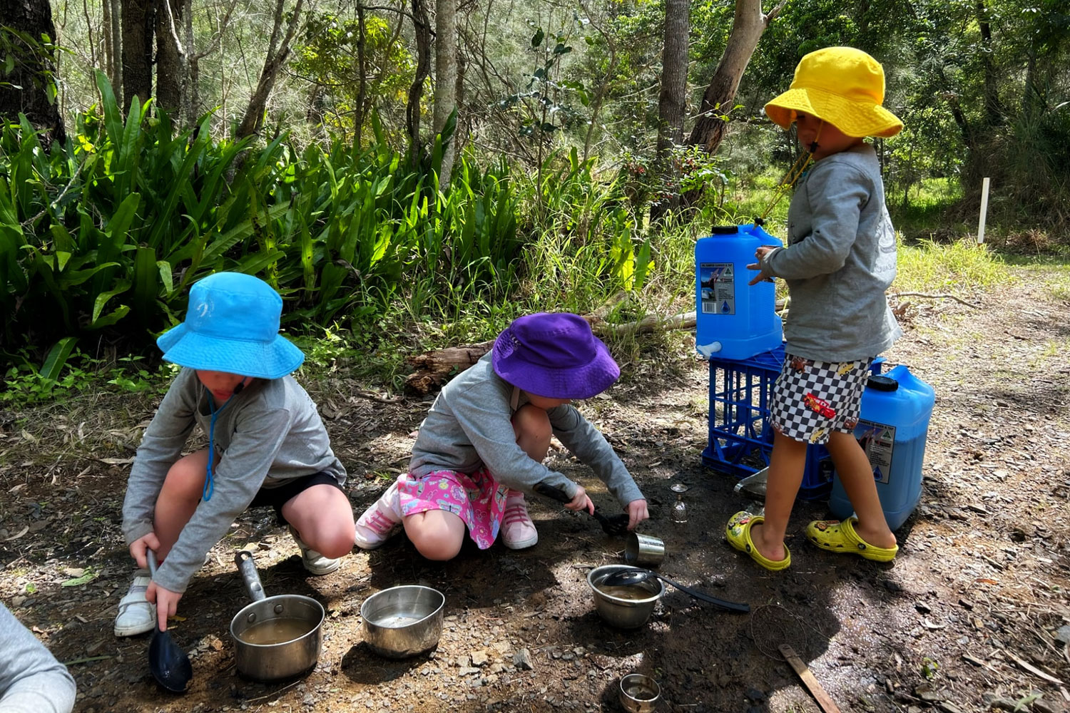 Three children are playing with water, saucepans and other utensils in the bush. They are wearing long sleeve shirts and wide brim hats